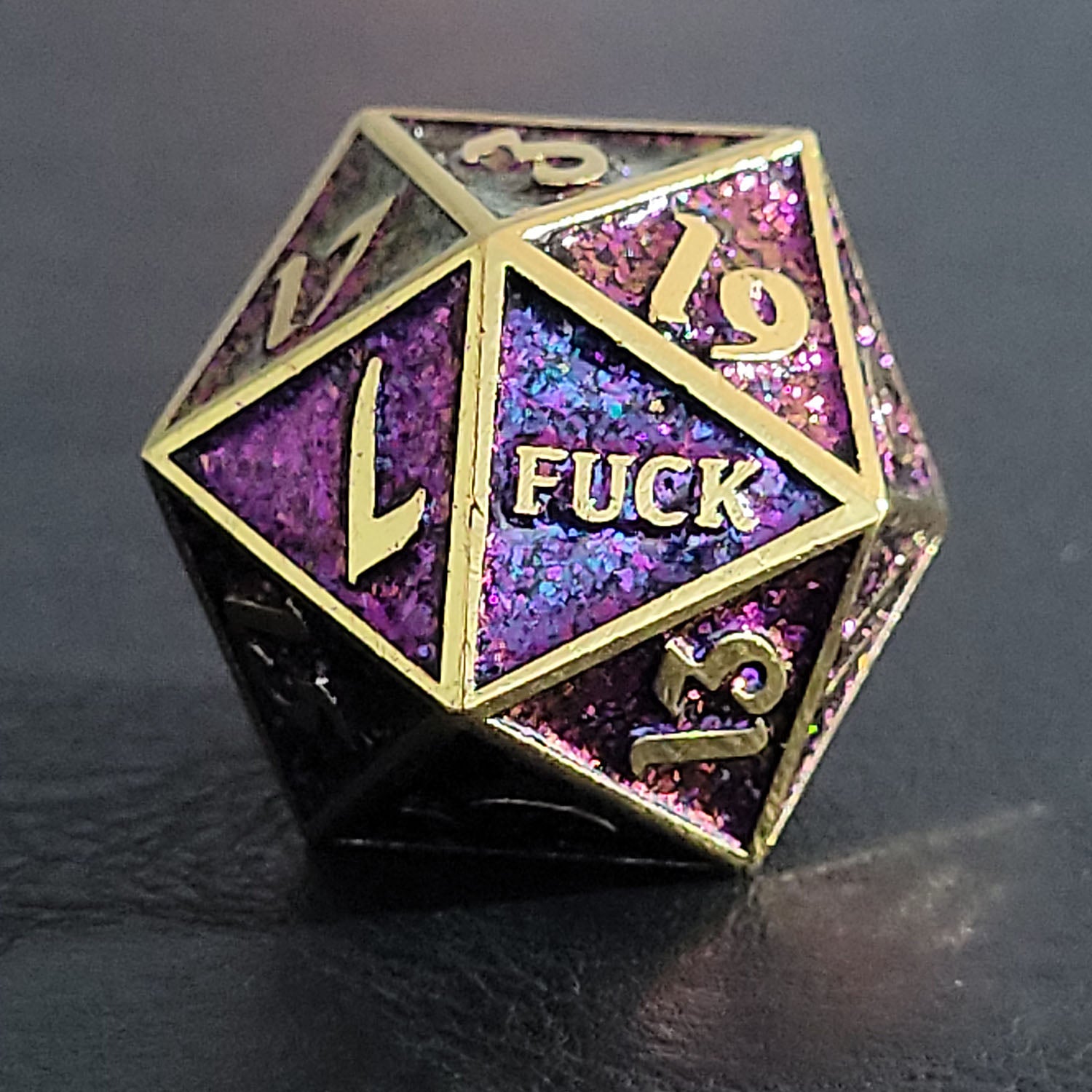 This is a close-up image of the Forged Gaming Gold Purple Glitter Fuck Yeah metal RPG D20 displaying the "Fuck" face.