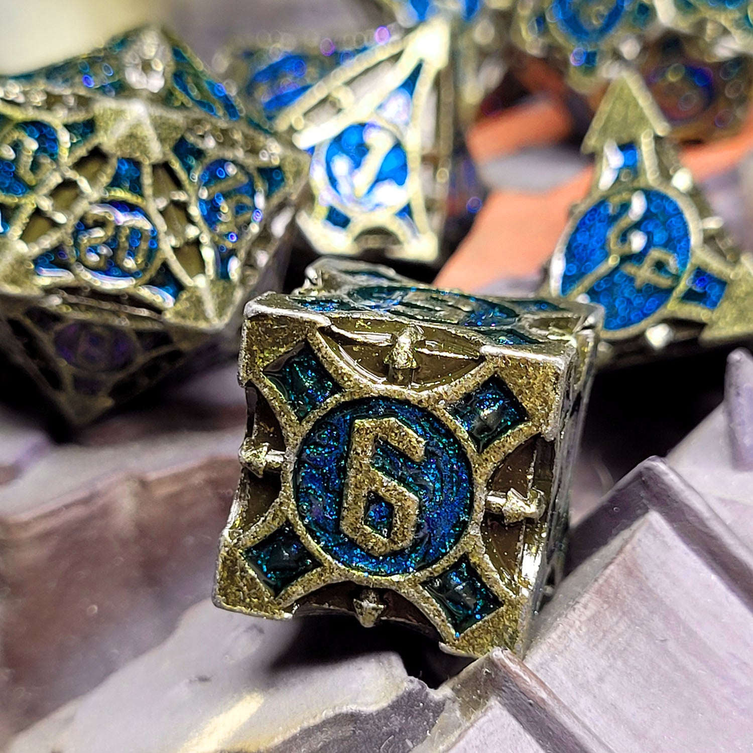 This is an environmental image of the Forged Gaming Arcane Enigma Gold Blue Metal RPG Dice Set. The photo was taken against the backdrop of a piece of wargaming terrain.