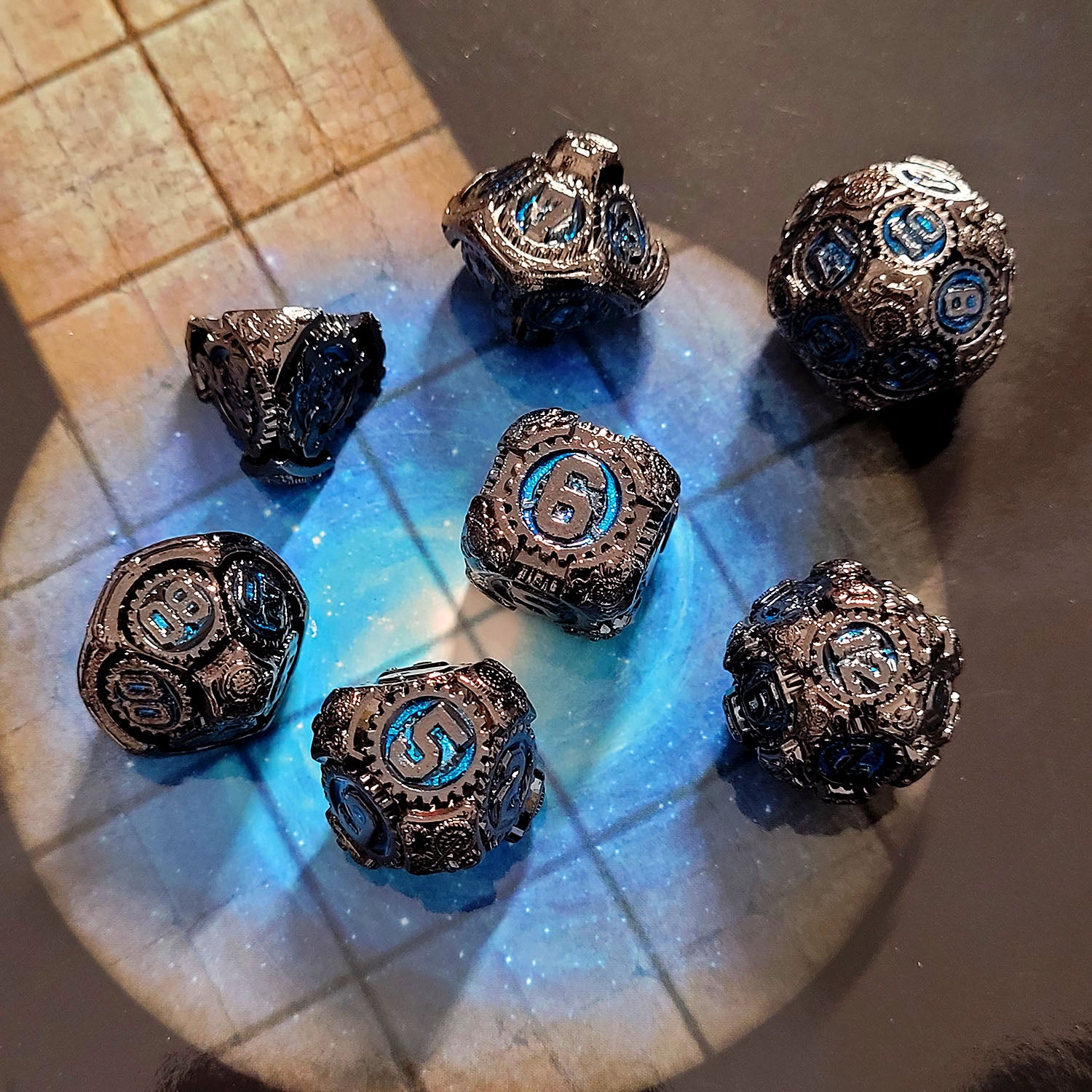 This is an environmental image of the Forged Gaming Chronomica Mechanicum Gunmetal Blue Metal RPG Dice Set. This photo was taken using a game board as a background.