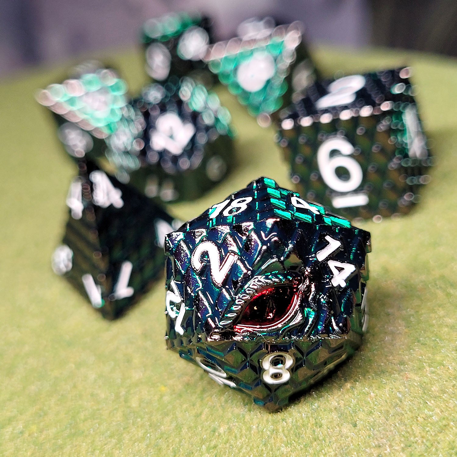 This is an environmental image of the Forged Gaming Dragon's Gaze Green Metal RPG Dice Set featuring red dragon eye on the D20 face. The image was taken using a piece of grass covered wargaming terrain as a background.