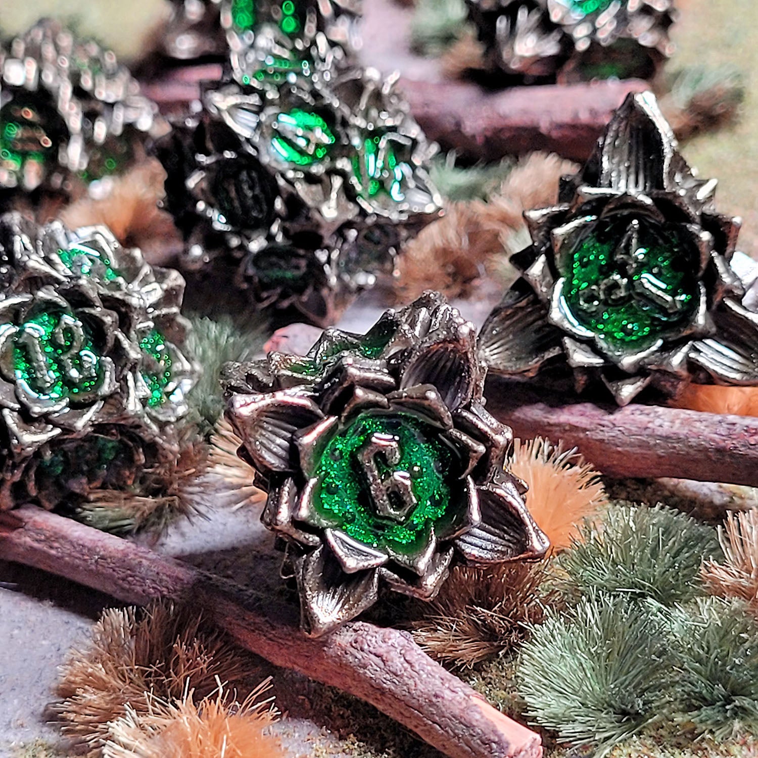 This is an environmental image of the Gothic Bouquet Antique Gold Green Metal RPG Dice Set. The photo was taken against a piece wooded clearing wargaming terrain. 