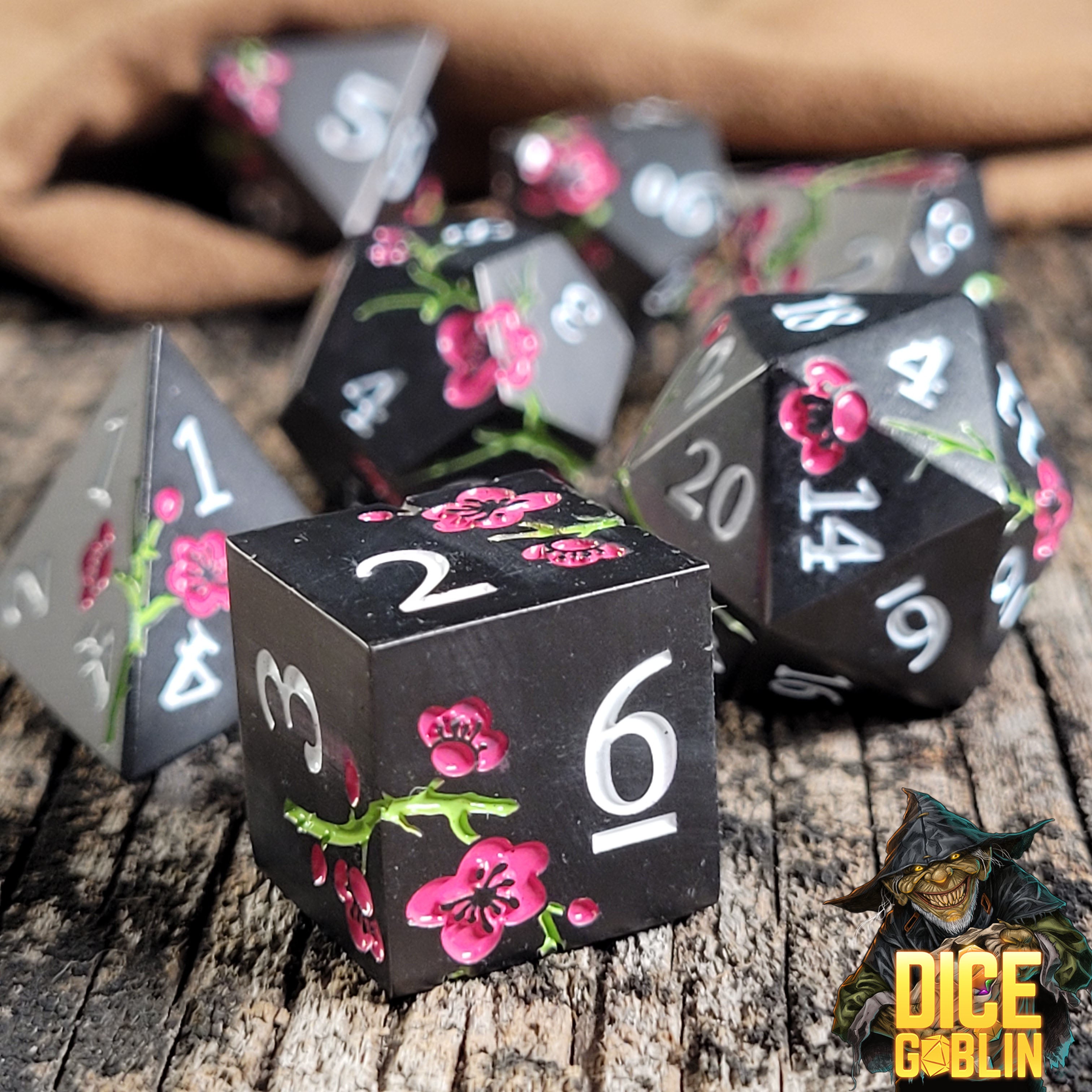 This is an environmental image of the Dice Goblin Flowers Black with Pink and Green Metal RPG Dice Set on a distressed wood surface.