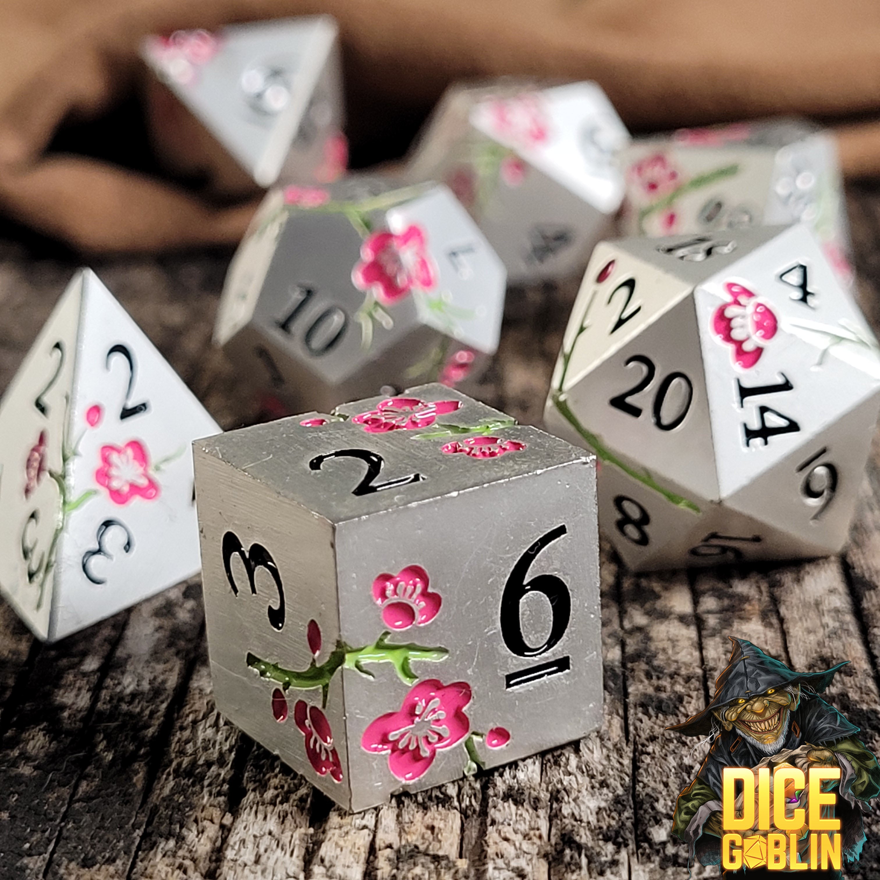 This is an environmental image of the Dice Goblin Flowers Silver with Pink and Green Metal RPG Dice Set on a distressed wood surface.
