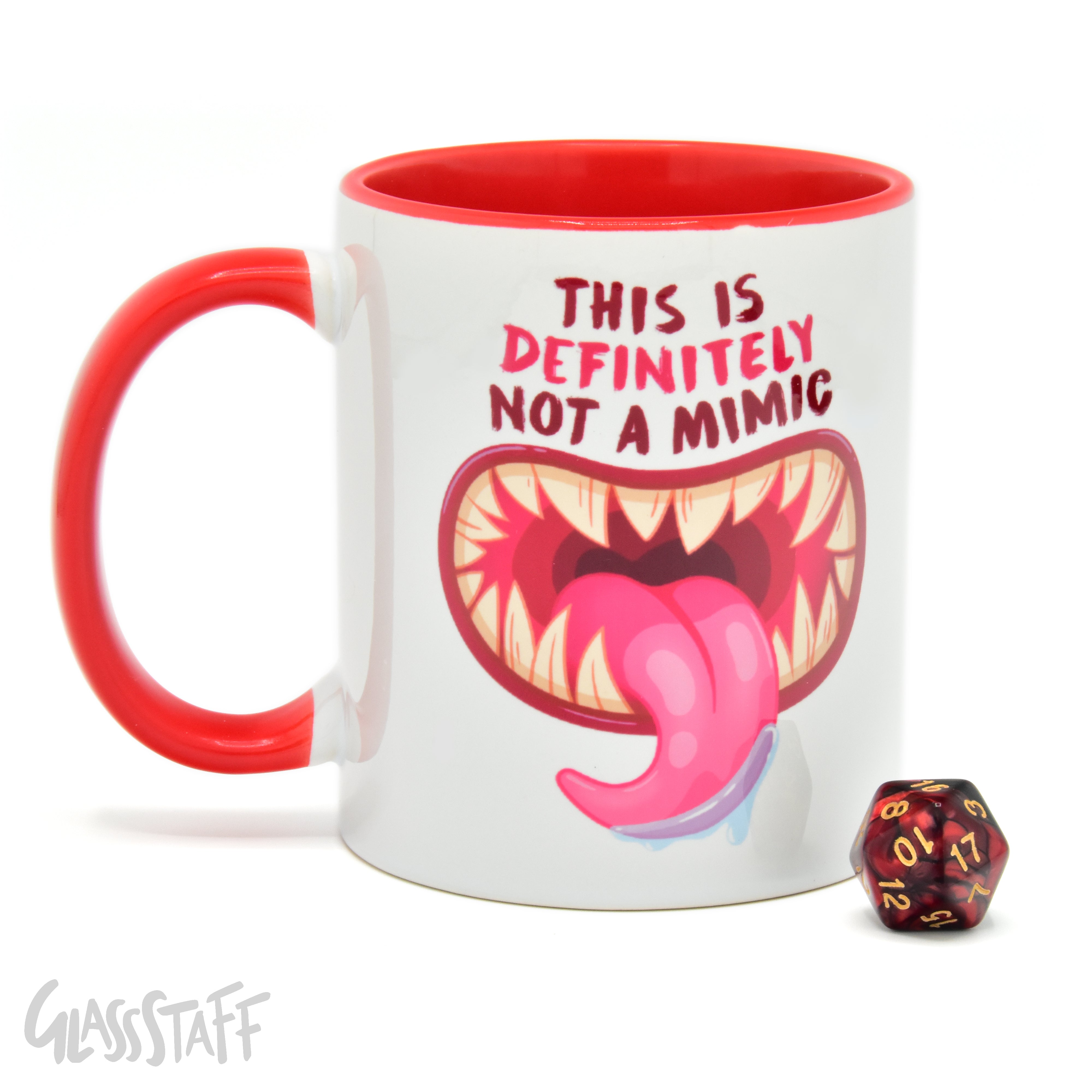 This is a white background image of the GLassStaff Not a Mimic ceramic mug. It shows the white mug with a red handle, red inside, and Not a Mimic design. It also shows the D20 die included with the mug.